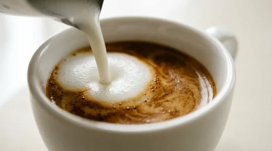 Close-up of velvety microfoam being poured into espresso creating glossy surface in ceramic cup