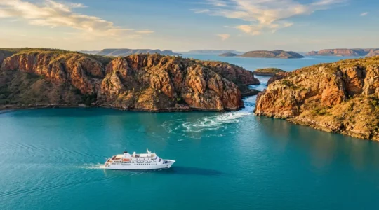 Luxury expedition cruise ship navigating turquoise waters near dramatic red cliffs and tidal waterfalls in Australia's remote Kimberley coast