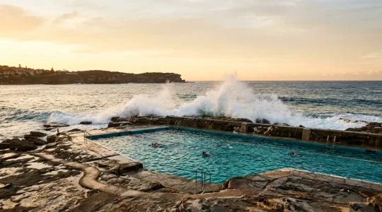 A serene Sydney ocean pool carved into coastal rocks with turquoise saltwater and dramatic wave spray at the edges during bluebottle season
