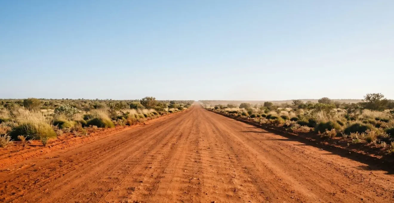 Wide environmental shot of an empty red dirt road stretching toward the horizon through Aboriginal land in Central Australia, with desert scrubland on both sides and vast open sky emphasizing the scale and remoteness