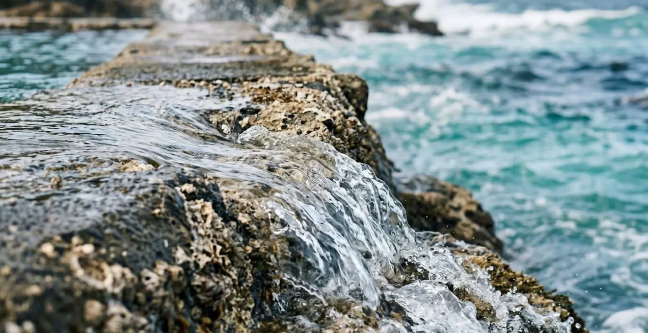 Close-up of turbulent ocean water flowing over a rocky pool wall edge during high tide showing natural pool flushing action